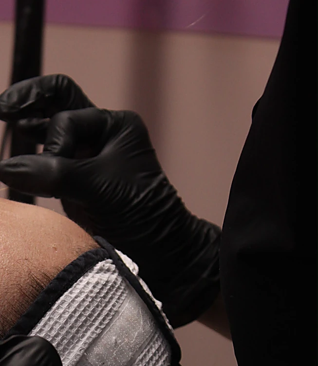 Manos con guantes negros trabajando el diseño de cejas con hilo, junto a la banda blanca de la cabina de The Brow + Lashes Bar.