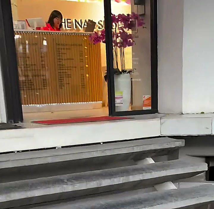 Entrada de The Nail Shop: escalones de concreto que suben a una puerta de vidrio con un tapete rojo y la recepción al fondo.