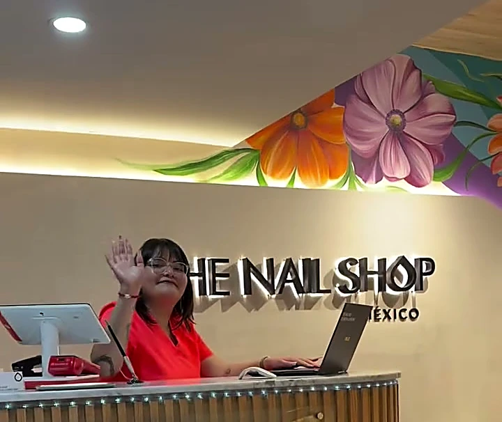 Recepción con el letrero de The Nail Shop, un mural de flores grandes en el techo y una recepcionista saludando.