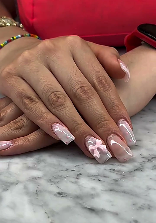 Uñas cuadradas largas en rosa translúcido con un moño blanco pintado, la mano apoyada en un cojín rojo.